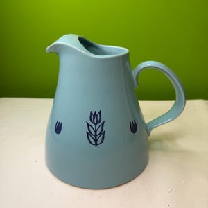 Può includere: Una brocca in ceramica azzurra con manico e beccuccio curvi. La brocca presenta un motivo floreale blu scuro. La brocca viene probabilmente utilizzata per servire bevande e ha una capacità di circa 1 litro.