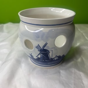 May include: A white ceramic pot with a blue windmill design. The pot has a wide opening, a blue rim, and two large circular holes. The design features a windmill, houses, and water, all in blue.