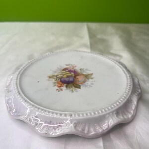 May include: White ceramic plate with a scalloped edge and a beaded border. The center of the plate features a colorful floral and fruit design. The plate is likely vintage and has a decorative purpose.