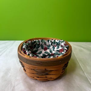 Peut inclure: Un petit panier rond et tressé avec une doublure en tissu décoratif présentant un motif de feuilles vertes et de baies rouges. Le panier est en bois brun clair avec une bordure marron plus foncée.
