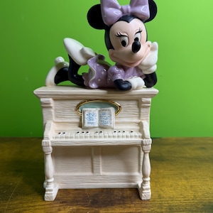 Peut inclure: Une figurine en c&eacute;ramique de Minnie Mouse portant une robe et un n&oelig;ud &agrave; pois lavande, posant son menton sur sa main tout en &eacute;tant allong&eacute;e sur un piano droit blanc cass&eacute;. Le piano pr&eacute;sente un pupitre dor&eacute; avec un livre de partitions ouvert.