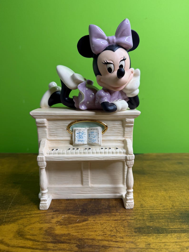 Peut inclure: Une figurine en c&eacute;ramique de Minnie Mouse portant une robe et un n&oelig;ud &agrave; pois lavande, posant son menton sur sa main tout en &eacute;tant allong&eacute;e sur un piano droit blanc cass&eacute;. Le piano pr&eacute;sente un pupitre dor&eacute; avec un livre de partitions ouvert.