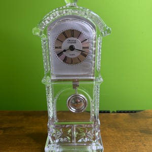 May include: A clear, decorative Crystal Legends quartz clock. The clock has a rectangular base and a domed top with a small, round finial. The clock face is silver with black hands and Roman numerals. A pendulum hangs below the clock face.