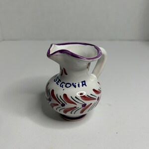 May include: A small white ceramic pitcher with a purple rim and handle. It has a red and purple floral design and the word "Segovia" written in blue.