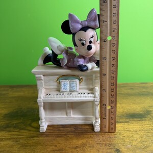 Peut inclure: Une figurine en c&eacute;ramique de Minnie Mouse portant un n&oelig;ud et une robe lavande, allong&eacute;e sur un piano droit de couleur cr&egrave;me. Une r&egrave;gle en bois plac&eacute;e &agrave; c&ocirc;t&eacute; de la figurine indique une hauteur totale d'environ 16,5 cm de la base au n&oelig;ud.