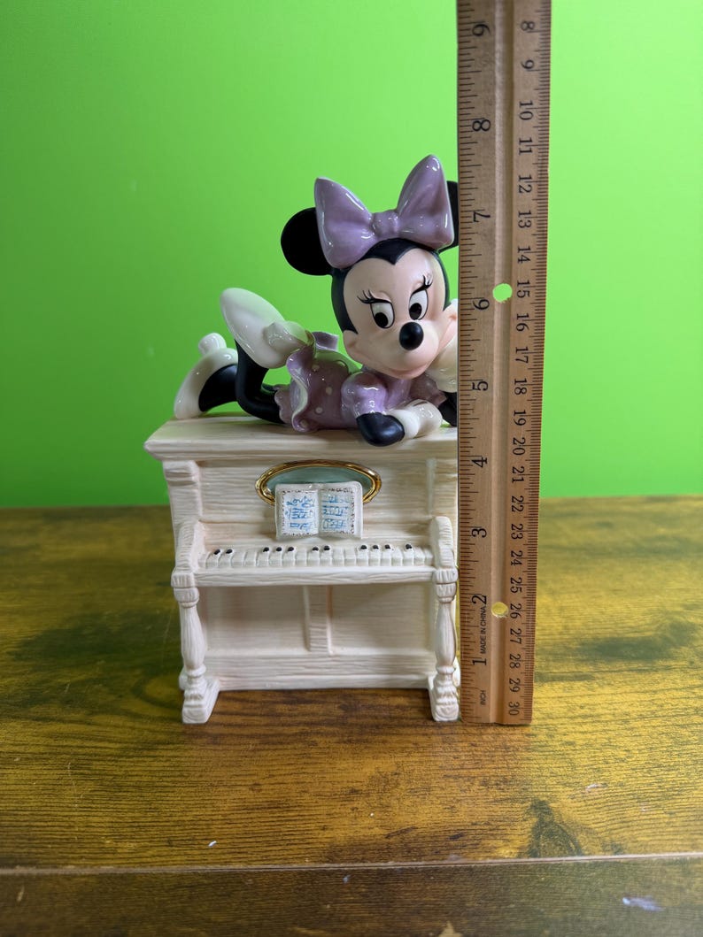 Peut inclure: Une figurine en c&eacute;ramique de Minnie Mouse portant un n&oelig;ud et une robe lavande, allong&eacute;e sur un piano droit de couleur cr&egrave;me. Une r&egrave;gle en bois plac&eacute;e &agrave; c&ocirc;t&eacute; de la figurine indique une hauteur totale d'environ 16,5 cm de la base au n&oelig;ud.