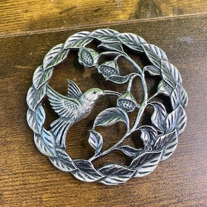 May include: A circular silver-toned metal trivet featuring an embossed design of a hummingbird hovering near a flowering branch. The outer border consists of a continuous wreath of detailed leaves, creating a decorative and functional kitchen accessory.