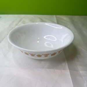 Puede incluir: Un cuenco de cerámica blanco con un patrón floral dorado alrededor del borde. El cuenco tiene una forma simple y redondeada y está sobre un fondo claro.