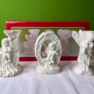 May include: White ceramic candle holders shaped like the letters J, O, and Y, each featuring a cherub angel design. The holders are displayed on a red box with white lettering.