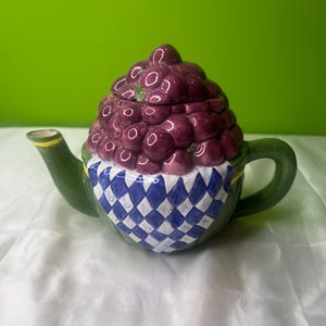 May include: A green ceramic teapot with a purple grape design. The teapot has a blue and white checkered pattern around the base.