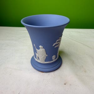 May include: A blue vase with white relief figures and a flared top. The vase features a round base and a detailed design of classical figures and trees. The vase is made of ceramic or porcelain.