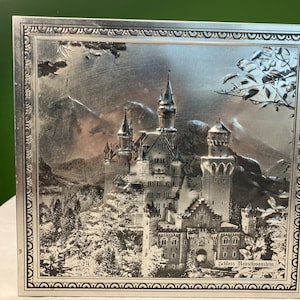 Puede incluir: Una caja de metal plateada con una imagen en relieve del Castillo de Neuschwanstein, un castillo de cuento de hadas con torres y torretas, sobre un fondo de montañas y árboles. La caja tiene un borde decorativo y el texto "Schloss Neuschwanstein" está impreso en la parte inferior.