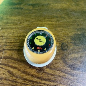 May include: A vintage Mampo Mile Meter, a small, round device with a yellow body and a white base. The meter has a black dial with white numbers and the text "Mampo Meter" in the center. The dial reads up to 9 miles.