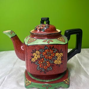 Peut inclure: Théière décorative en céramique rouge avec une poignée et un couvercle noirs. Elle présente des motifs floraux jaunes, oranges et bleus, avec des accents verts. Posée sur un socle assorti. Un service à thé vintage.
