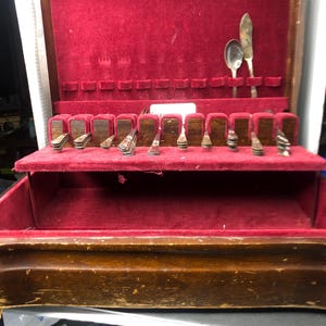 Puede incluir: Un estuche de cubiertos de madera abierto con interior de terciopelo rojo. El estuche contiene varias piezas de cubertería, incluyendo cucharas, cuchillos y tenedores. La cubertería está dispuesta en ranuras designadas dentro del estuche.