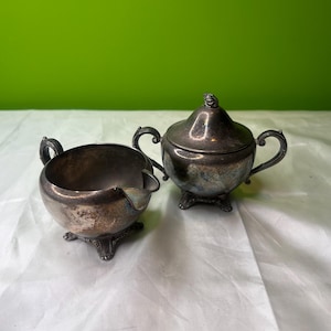 Puede incluir: Juego de azucarero con tapa y jarrita para crema plateados antiguos. El azucarero tiene forma redondeada con tapa y un pequeño asa decorativa. La jarrita tiene un pico y un asa. Ambas piezas tienen pies ornamentados.