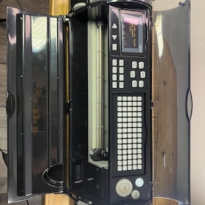 Puede incluir: Una máquina Cricut negra y gris con la tapa abierta, que revela una pantalla y un teclado. La pantalla muestra texto. La máquina tiene un rodillo plateado y una serie de botones. La máquina está sobre una superficie de madera.