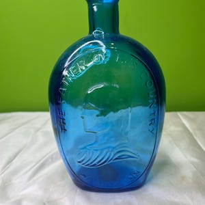 Puede incluir: Una botella de vidrio azul y verde vintage con forma redondeada. La botella presenta una imagen en relieve de un perfil y las palabras "THE FATHER OF HIS COUNTRY" y "WASHINGTON".