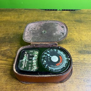 May include: A vintage Weston Master II universal exposure meter housed in a brown leather case with a hinged lid. The device features a black circular dial with white numerical scales, a central silver disc, and a rectangular window with text.