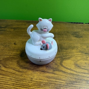 May include: A white cat-shaped kitchen timer with a gray base and a small gray mouse figurine. The timer has a dial with numbers and a pink nose on the cat. The timer is on a wooden surface.