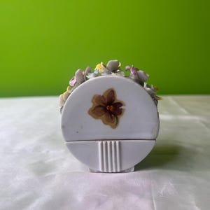 Könnte beinhalten: Eine weiße Porzellan-Schmuckdose mit Blumenmuster. Die Dose ist halbrund und hat eine einzelne Blume, die auf die Vorderseite gemalt ist.