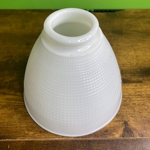 Puede incluir: Una pantalla de lámpara de vidrio blanco con una silueta en forma de campana y un patrón de cuadrícula texturizado en la superficie exterior. La pantalla presenta una abertura circular lisa en la parte superior y una base abierta más ancha.