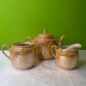 Puede incluir: Un juego de té de cerámica marrón con tetera, jarra de crema y azucarero. El juego tiene un acabado brillante y un sutil brillo iridiscente.