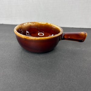 Könnte beinhalten: Eine kleine braune Keramikschüssel mit Henkel. Die Schüssel hat eine glasierte Oberfläche und eine braune und beigefarbene Farbgebung.