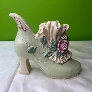 Puede incluir: Un zapato decorativo de cerámica en un tono verde suave, con tacón alto y adornado con una rosa rosa y detalles con volantes. El zapato tiene una estética vintage, con detalles delicados y un motivo floral.