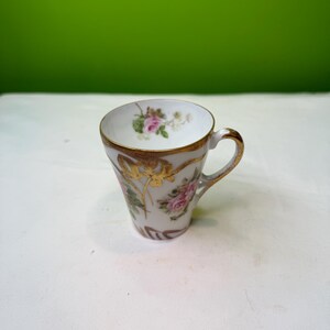 May include: A small, white teacup with a gold rim and handle. The cup is decorated with pink roses and gold floral accents. The teacup is likely vintage or antique.