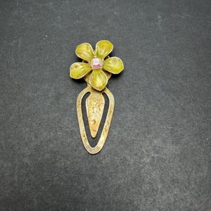 May include: A metal bookmark featuring a five-petaled yellow flower at the top with a central iridescent pink rhinestone. The lower portion of the bookmark is a teardrop-shaped loop with a textured, weathered finish in a muted gold tone.