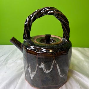 Op de afbeelding: Een donkerbruine keramische theepot met een gedraaide handgreep. De theepot heeft een geometrische vorm met witte golfpatronen rond de basis. Het deksel heeft een kleine, donkere knop.