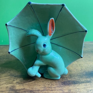 May include: A ceramic figurine of a light green rabbit standing under a matching light green umbrella. The rabbit has dark blue eyes and an orange interior on one ear. The umbrella features thin black lines radiating from the centre to the edges.