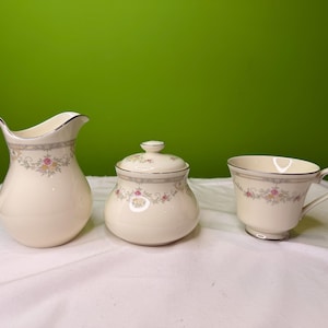 May include: A cream-colored porcelain tea set with floral and silver trim. The set includes a creamer, a covered sugar bowl, and a teacup. The floral design features pink and yellow flowers.