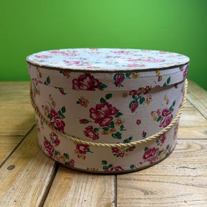 May include: A round hat box with a floral pattern of red roses on a cream background. It features a gold rope handle and trim. The box is on a wooden surface.