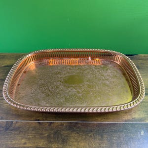 Puede incluir: Una bandeja de servir rectangular de color cobre con un patrón floral decorativo en el centro. La bandeja tiene un borde en forma de cuerda y un borde perforado. La bandeja está sobre una superficie de madera con un fondo verde.