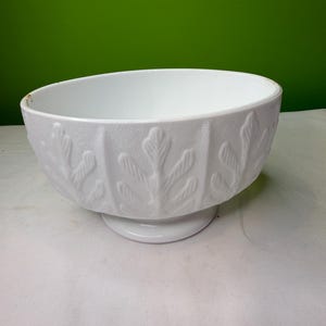 Peut inclure: Bol en céramique blanc avec un socle. Le bol présente un motif de feuilles texturées sur l'extérieur. Le bol est posé sur une surface blanche et un fond vert.