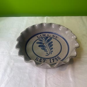 Puede incluir: Un plato para tartas de cerámica blanca con un diseño azul y blanco. El plato tiene un borde festoneado y presenta el texto "LIMA, NY EST 1788" y un diseño de hoja estilizado.