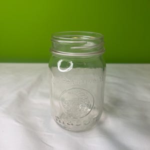 Puede incluir: Un tarro de cristal transparente con boca ancha y tapa. El tarro tiene un logotipo verde de cosecha con una imagen estilizada de una fruta y la palabra "Mason" impresa en la parte inferior.