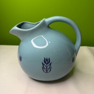 Può includere: Una brocca in ceramica azzurra con un corpo arrotondato e un manico curvo. La brocca è dotata di un beccuccio per versare ed è decorata con disegni floreali blu scuro. La brocca è adatta per servire bevande.