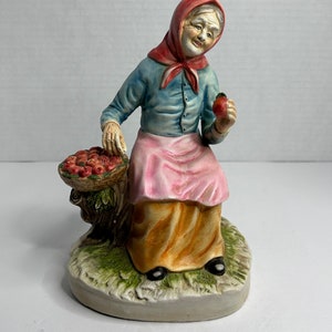 Könnte beinhalten: Eine Keramikfigur einer älteren Frau, die ein rotes Kopftuch und ein blaues Hemd trägt. Sie sitzt auf einem Baumstumpf und hält einen roten Apfel in der Hand. Sie hat einen Korb mit roten Früchten auf dem Schoß.