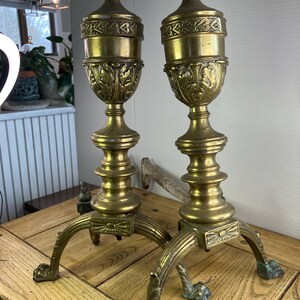 May include: Two antique brass candle holders. Each holder has a decorative urn-shaped top with leaf and floral detailing. The base features three curved legs ending in claw feet. The candle holders are displayed on a wooden surface.