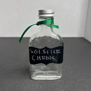 May include: A small clear glass bottle with a silver lid and a green ribbon tied around the neck. The bottle has a black label with the words "SOLSTICE CHEER" written in white.
