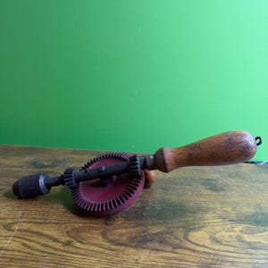 May include: A vintage hand drill with a wooden handle and a red gear. The drill has a black chuck and a metal frame. The tool is designed for manual drilling and is likely used for woodworking or other crafts.