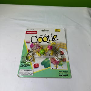 Può includere: Un gioco portachiavi Cootie, confezionato su una carta verde e bianca. La confezione include miniature di Cootie e un tappetino di gioco. Il testo "Cootie" è ben visibile, con un avvertimento sulle piccole parti.