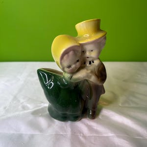 May include: A vintage ceramic planter shaped like a couple holding each other. The woman is wearing a yellow hat and dress, and the man is wearing a brown hat and jacket. The planter is green and has a leaf-shaped base.