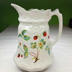 May include: White ceramic pitcher with a green, red, and yellow floral design featuring strawberries and flowers. The pitcher has a scalloped rim and a handle.