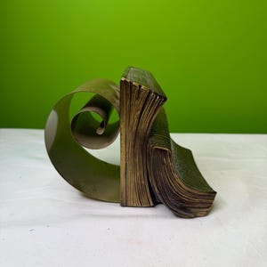 May include: A bronze-colored bookend set with a unique design. One bookend is a stack of pages, and the other is a curled, ribbon-like shape. The bookends are on a white surface with a green background.