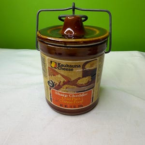 May include: A brown ceramic cheese crock with a lid and metal handle. The label reads "Kaukauna Cheese Sharp Cheddar Cold Pack Cheese Food" and "NET WT. 12 OZ 340g". The crock is cylindrical and sits on a white surface against a green background.