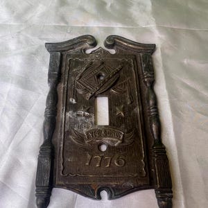 Puede incluir: Una cubierta de interruptor de luz vintage con un diseño decorativo que presenta una flauta y un tambor, estrellas y el texto "Fife & Drum 1776".